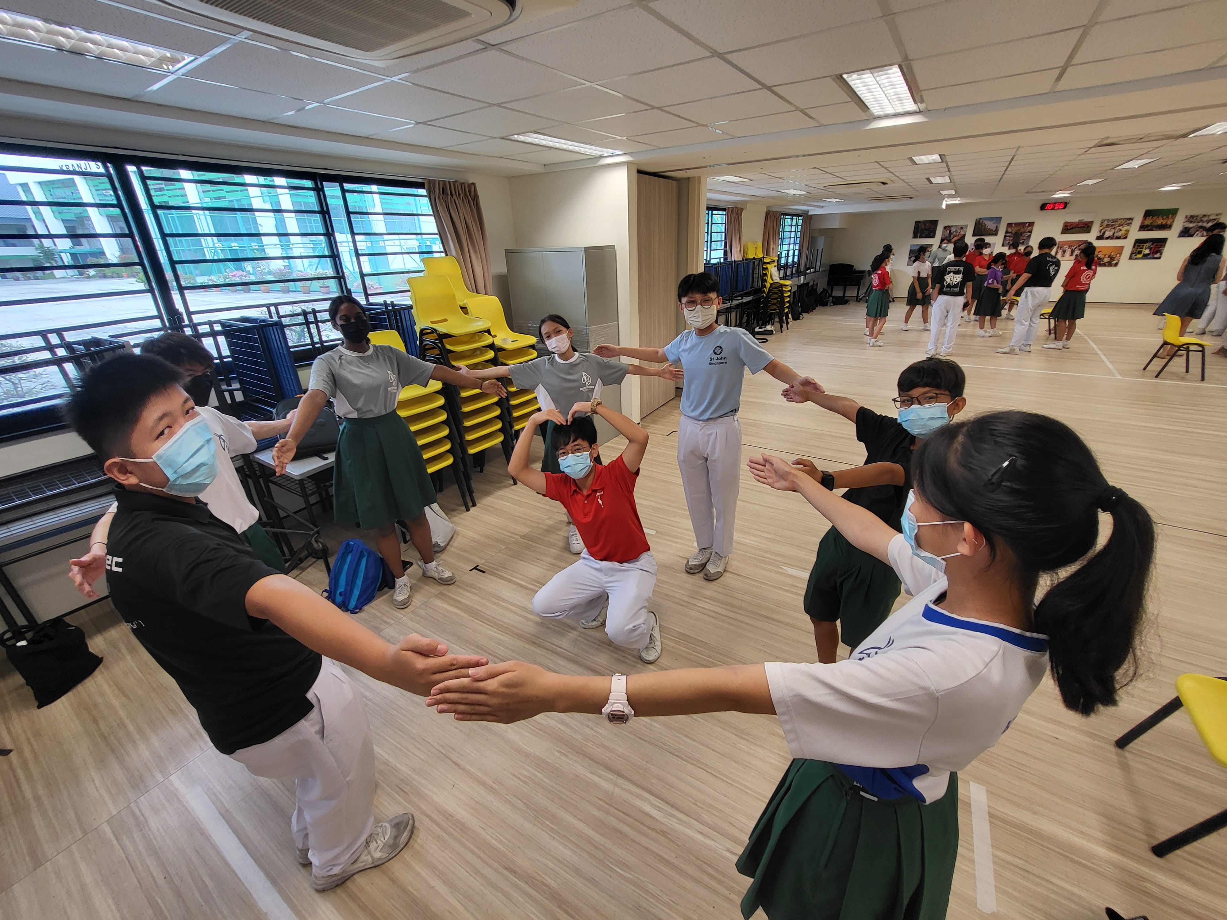Students wearing masks form a circle with outstretched arms in a bright school activity room.